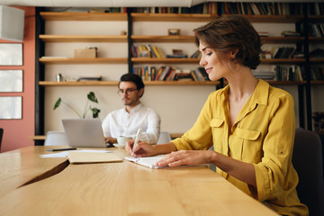 Young beautiful woman sitting at the table happily writing in notepad with colleague on background at work in office