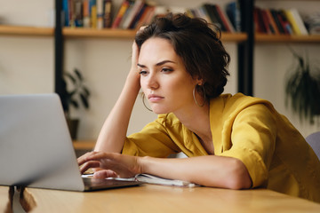 Young upset woman sitting at the desk tiredly working on new project with laptop in office
