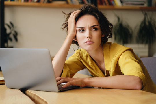 Portrait of young serious businesswoman sitting at the desk tiredly looking aside while working on laptop in modern office