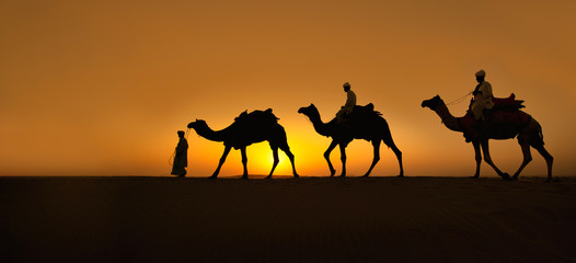 Fototapeta na wymiar Rajasthan travel background - Three indian cameleers (camel drivers) with camels silhouettes in dunes of Thar desert on sunset. Jaisalmer, Rajasthan, India