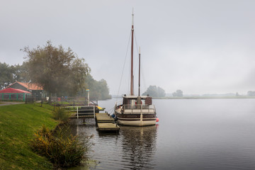 Fototapeta na wymiar Behind a lock to the North Sea is this channel and lake near Emden. A sailboat is located on the jetty in front of a restaurant in fog and mist. It's a morning in the fall.