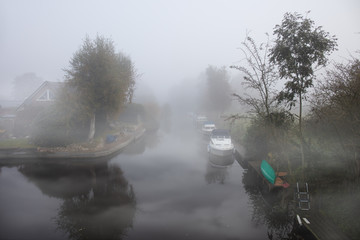 A channel on the big sea in northern Germany near the port city of Emden in heavy fog in the early morning. A house on the canal with many boats can be seen. It is autumn.
