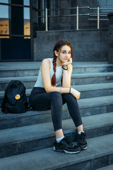 Beautiful European student with a backpack sitting on the steps near the University, close-up. A young pretty student in jeans and sneakers resting after class.