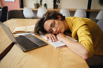 Young tired woman in eyeglasses asleep on desk with laptop and notepad under head at workplace