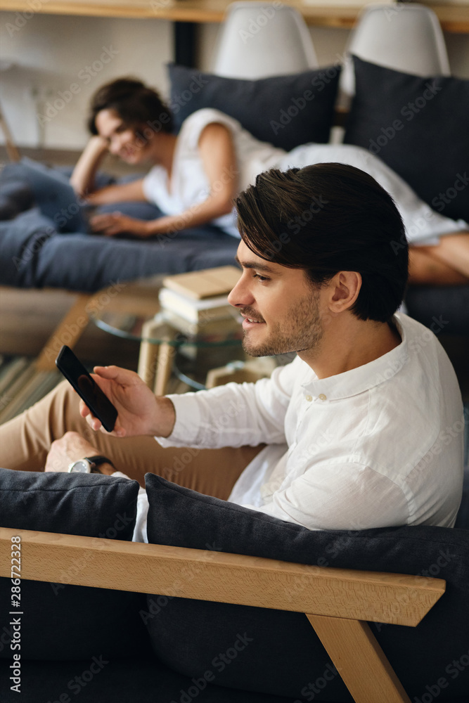 Aufkleber Side view of young smiling businessman sitting on sofa with cellphone happily looking aside in co-working space