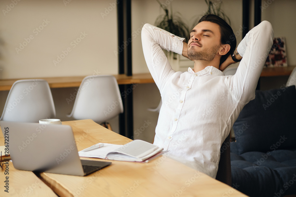 Poster young attractive man sitting at the table with laptop and notepad leaning on chair while dreamily re