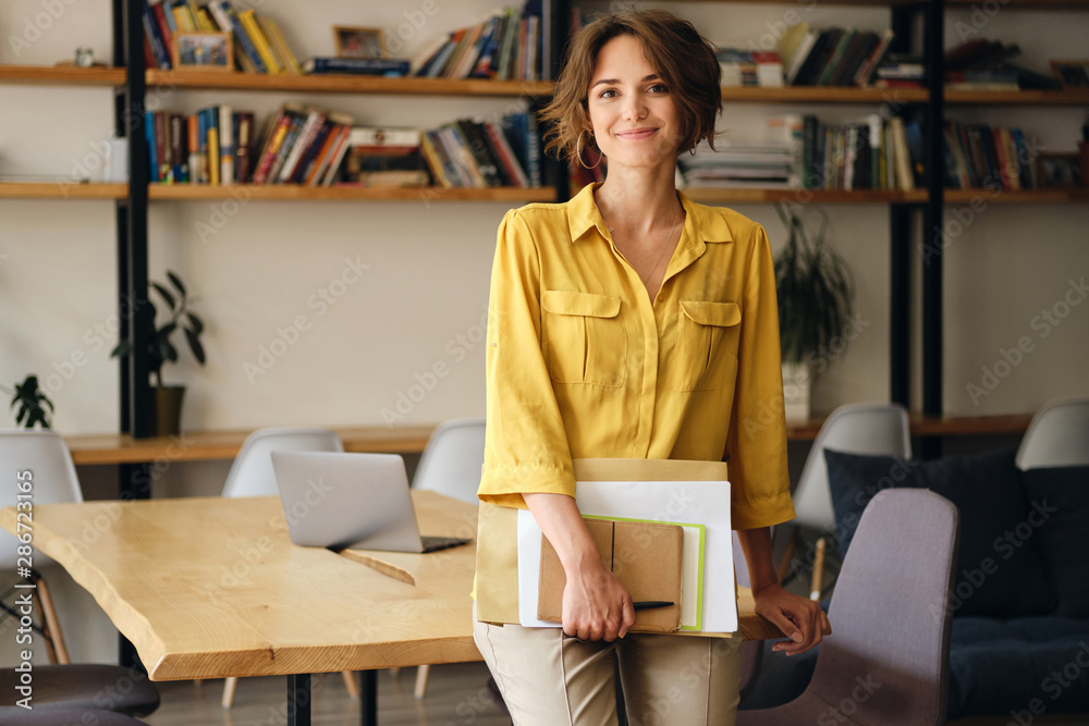 Fototapete Young beautiful woman in yellow shirt leaning on desk with notepad and papers in hand while happily looking in camera in modern office