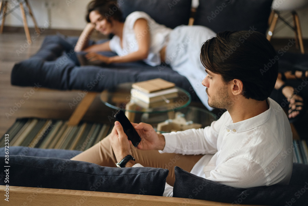 Poster Side view of young businessman sitting on sofa with colleague on background while thoughtfully using cellphone in co-working space