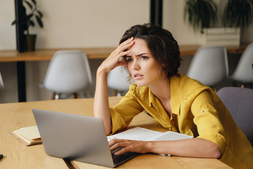 Young exhausted woman sitting at the desk with laptop while tiredly holding hand near head at work...