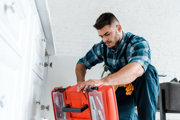 selective focus of handsome handyman looking at opened toolbox in kitchen