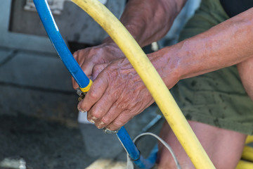 Alter Mann mit nassen Händen verbindet zwei Wasserschläuche draußen im Sommer, Deutschland