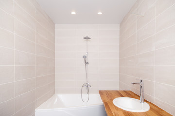 Empty new bathroom with beige ceramic rectangular tiles, large bathtub, silver shower, water tap, wooden worktop with ceramic sink. Concept repair bathroom, renovation in apartments, hotel