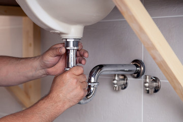 Closeup male plumber worker in blue denim uniform, overalls, fixing sink in bathroom with tile wall. Professional plumbing repair service, installation water pipes, man mounted sewer drain