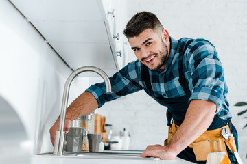 selective focus of happy handyman working near sink in kitchen - obrazy, fototapety, plakaty