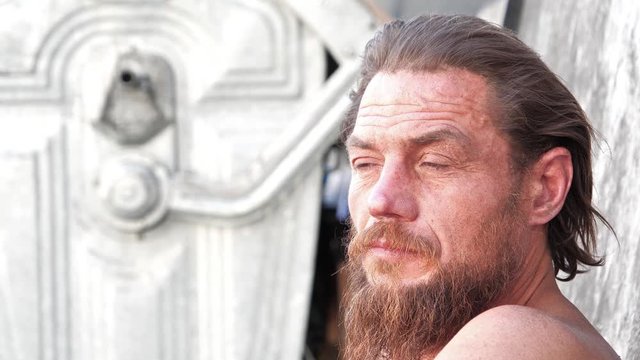 Down-and-out person sits near refuse bin. Real person with no fixed abode, homeless man