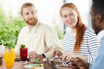 Group of young friends sitting at the table eating vegetable salad and talking to each other during lunch in cafe