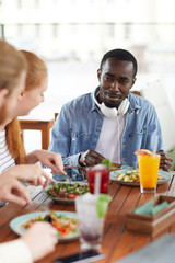 Fototapeta na wymiar Young African man sitting at the table eating salad and talking to his friends they have lunch at restaurant