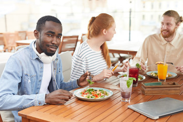 Fototapeta na wymiar African young man eating vegetable salad and smiling at camera while sitting at the table with his friends during lunch in cafe