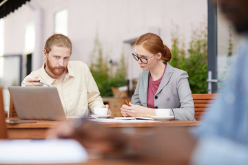 Young bearded businessman pointing at laptop computer and discussing online project with young businesswoman in eyeglasses while they sitting in outdoor cafe