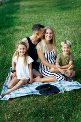 Fototapeta premium Picture of kissing parents with children sitting on plaid on picnic on summer day