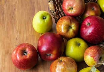 Red and green apples spilled out from a wicker basket lying on its side onto a brown wooden board, top side view
