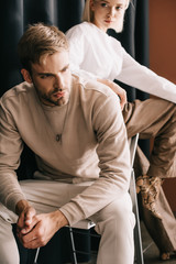 blonde woman in white blouse and bearded man sitting on chair near curtain