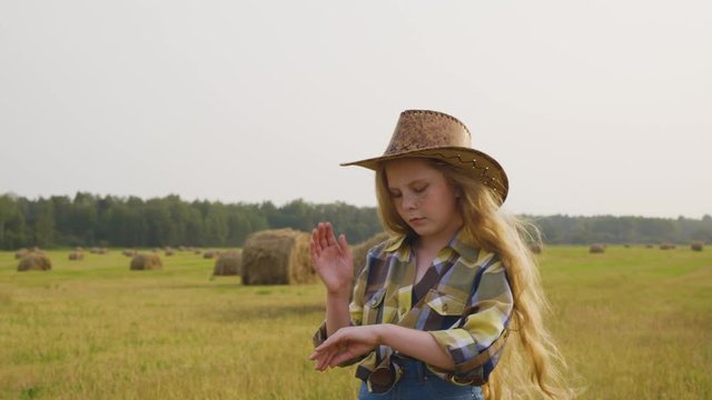 Cowgirl on countryside field on haystack background. Young cow girl in hat posing front camera. Country girl in checkered shirt on harvesting field in village