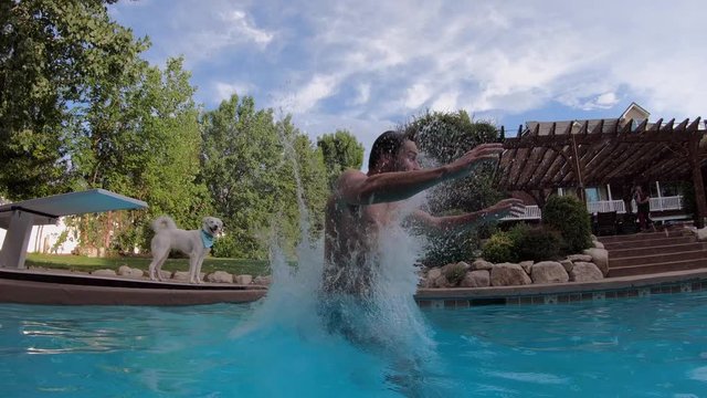 Man doing back flip gainer into pool as dog watches then walks away as he splashes.