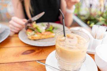 Focus in foreground on Cold coffee with ice cubes. A young woman is having Breakfast in a summer cafe.