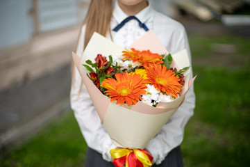 School girl dressed in school uniform holding a bright orange and yellow festive bouquet of beautiful flowers for teacher. No face. Blurred background.