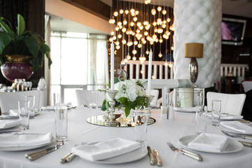 Wedding table setting is decorated with fresh flowers and white candles. Wedding floristry. Bouquet with roses, hydrangea and eustoma. On the background blur are burning garlands with light bulbs