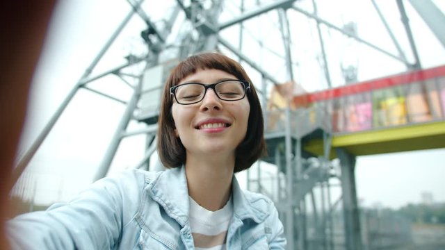Slow motion of attractive young woman making online video call near observation wheel in city on summer day standing alone talking gesturing waving hand.