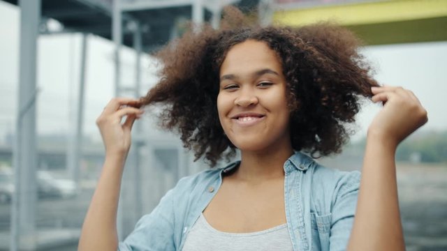 Portrait of playful African American girl messing hair then looking at camera smiling standing outdoors alone in city. People, lifestyle and hairstyle concept.