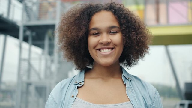 Slow motion of pretty African American girl smiling looking at camera outdoors standing alone with happy face. Youth, portrait and happiness concept.