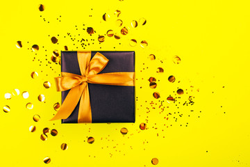 Black gift box with golden bow on yellow background with glitter. Holiday concept.
