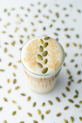 Milky latte with pumpkin seeds on froth, served on white background with spreaded pumpkin seeds