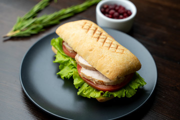 Close up of toasted ciabatta with tomatoes, smoked chicken breast and bunch of green leaves with olive oil and pepper, served on dark wood table with knife and fork