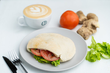 A cup of nice cappuccino and pita, stuffed with veggies and chicken breast on a white table in cafe
