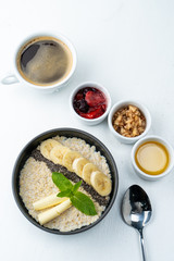 Healthy sweet breakfast with whole grain porridge, chia seeds, butter, banana and other toppings, served with a cup of black coffee on a white background