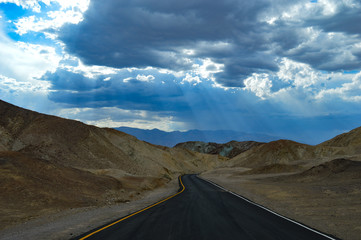 Sun rays shine through the clouds over the desert mountains and freshly-paved road of Death Valley National Park
