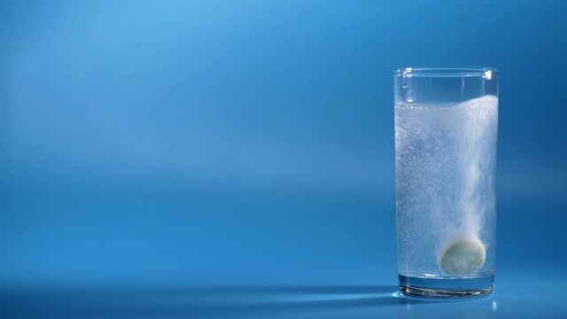 Effervescent Tablet Falling In Water. Pill Falls And Dissolves With Bubbles. Effervescent Tablet In Glass Of Water On Blue Background. Shot Of Effervescent Pill Dissolving In A Glass Of Water.