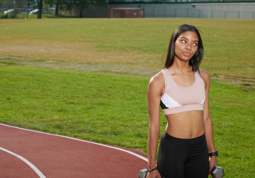 Stunning young black woman, fitness model, works out on outdoor track - playfully looking off frame while holding weights