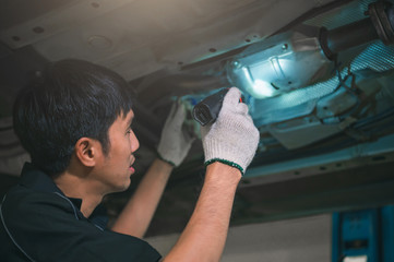 Asian male mechanical hold and shining flashlight to examine car under chassis of automotive...