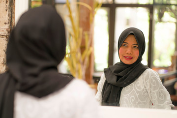 lifestyle portrait of elegant and happy middle aged Asian Indonesian Muslim woman in traditional hijab adjusting the Islamic scarf in front of the mirror