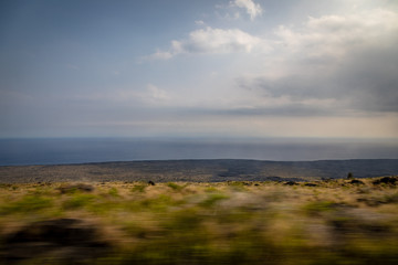 The foreground whizzes by looking out to a flat ocean fading into the horizon with a dark lava field and dry yellow and green bushes from a moving vehicle in Hawaii