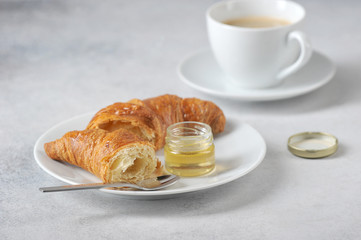 Croissant on a plate and a cup of coffee. Near the croissant a jar of honey. Close-up. Light background.