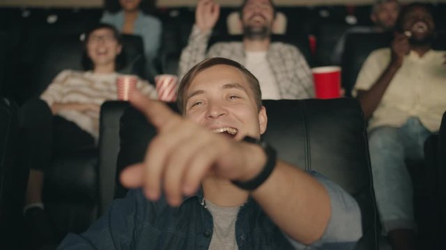 Happy young guy with short blond hair is laughing eating popcorn pointing at screen watching movie in cinema having fun. Joyful people and snacks concept.