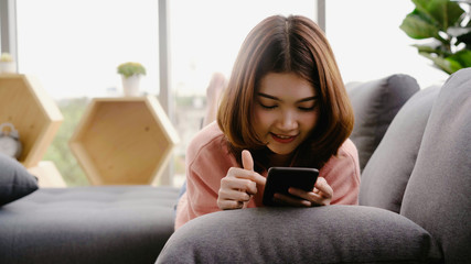 Portrait of beautiful attractive young smiling Asian woman using smartphone while lying on the sofa when relax in living room at home. Enjoying time lifestyle women at home concept.