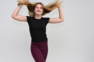 A knee-length portrait of a pretty brunette girl with long flowing hair in a black T-shirt and burgundy trousers on a white background. Smiling, showing emotions.
