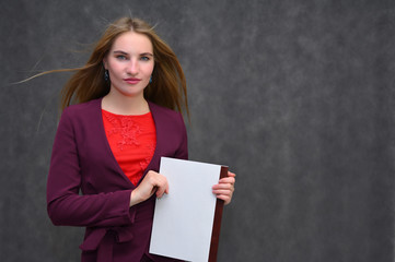 Portrait of a pretty secretary manager brunette girl with long flying hair in a burgundy business suit with a folder on a gray background. Smiling, showing emotions.